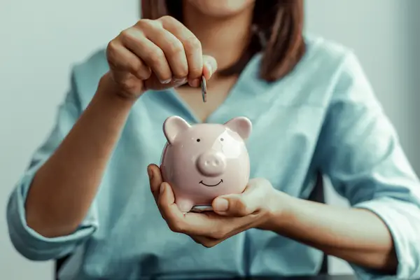 A person in a blue shirt, perhaps reminiscing about cherished May memories, carefully places a coin into a pink piggy bank.