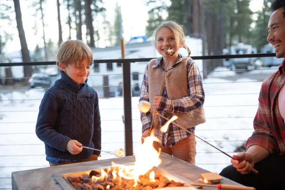 Two children and an adult toast marshmallows over an outdoor fire pit at Coachland RV Resort. Snow and trees create a picturesque backdrop, capturing the essence of Truckee-Tahoe's winter charm.