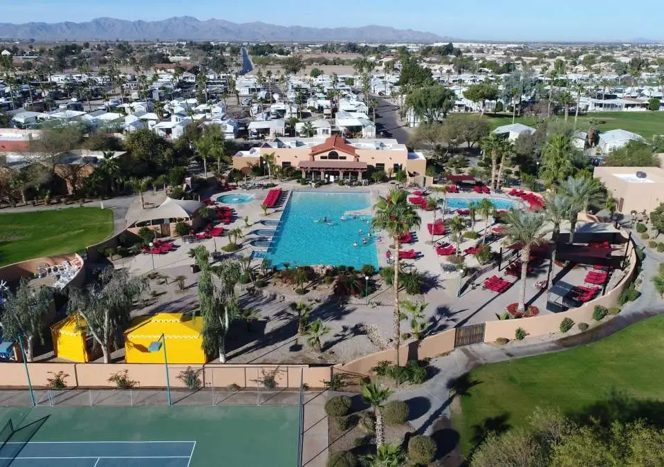 Aerial view of Pueblo El Mirage Resort in Arizona, showcasing a large swimming pool, red lounge chairs, palm trees, and a tennis court. Nestled in a vibrant retirement community, it offers stunning vistas of the surrounding residential area with majestic mountains in the background.