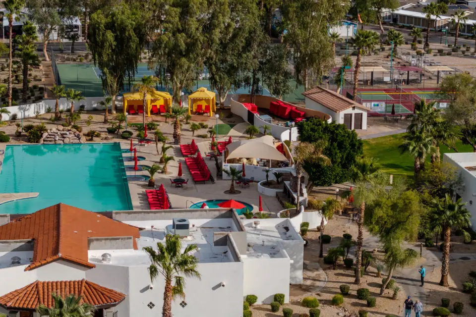 Aerial view of a resort with a swimming pool, red lounge chairs, and yellow cabanas. Tennis courts are nestled among trees and greenery, perfect for RVing enthusiasts to unwind after a day of adventure.