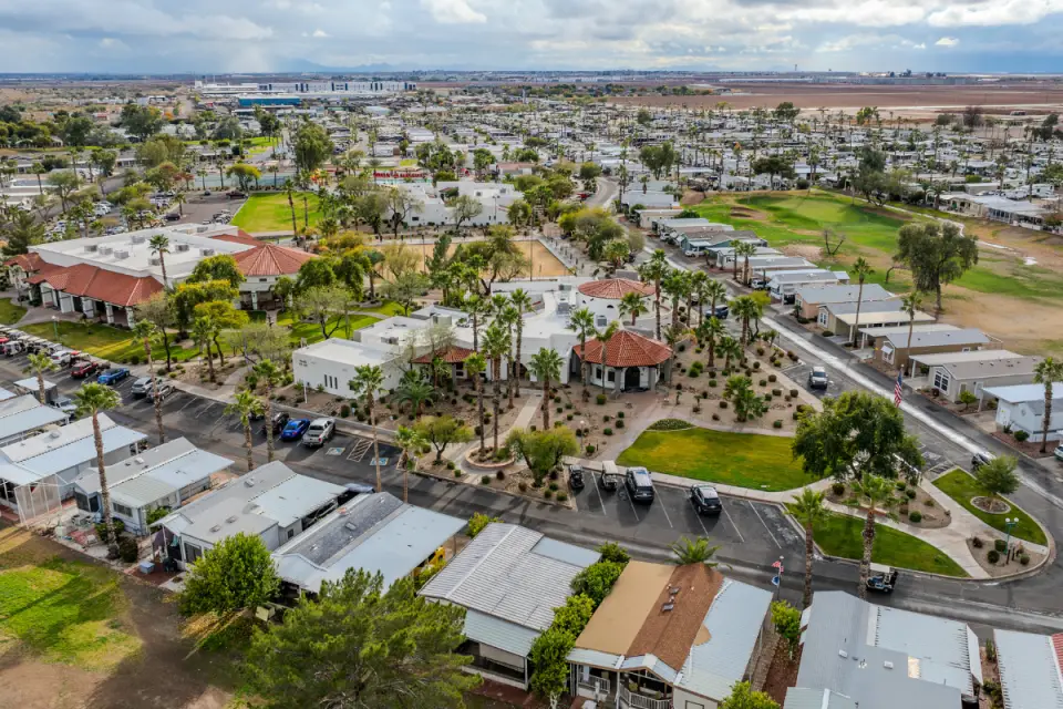 Aerial view of a residential community with houses, greenery, and roads. In the background, larger buildings and open land stretch under a cloudy sky. RVing enthusiasts might appreciate the scenic routes, while others see the benefits in securing a manufactured home here.