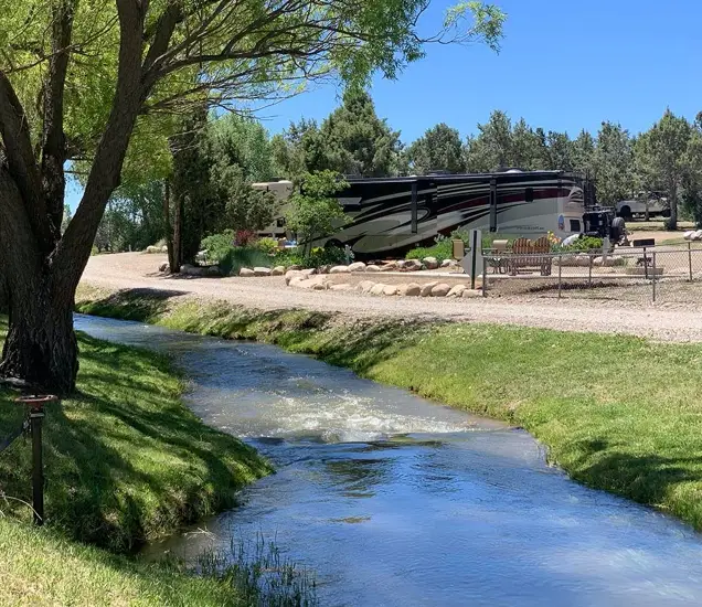An RV is parked beside a small stream with green grass and trees on a sunny day, perfect for a visit in the scenic beauty of Durango, Colorado.