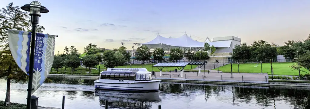 A small boat is docked on a tranquil Texas river beside a park with a white canopy structure and lush greenery under a clear sky, offering the perfect setting for fun-filled travel adventures.