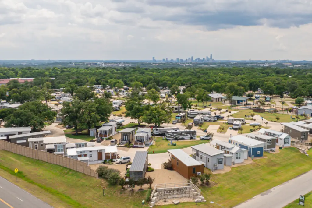 Aerial view of a mobile home park with various trailers and RVs, embraced by Austin's lush greenery. The distant city skyline peeks under a partly cloudy sky, perfect for those seeking insights into the RV lifestyle.
