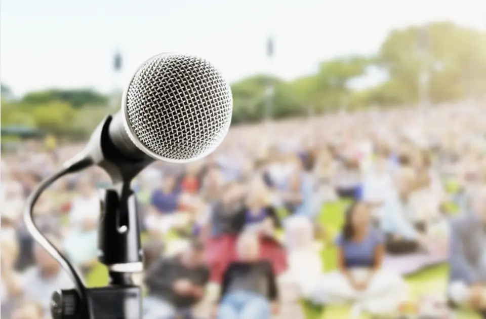 Close-up of a microphone on a stand with a blurred background showing a large crowd seated outdoors on a grassy area, basking in the golden hues of sunrise.
