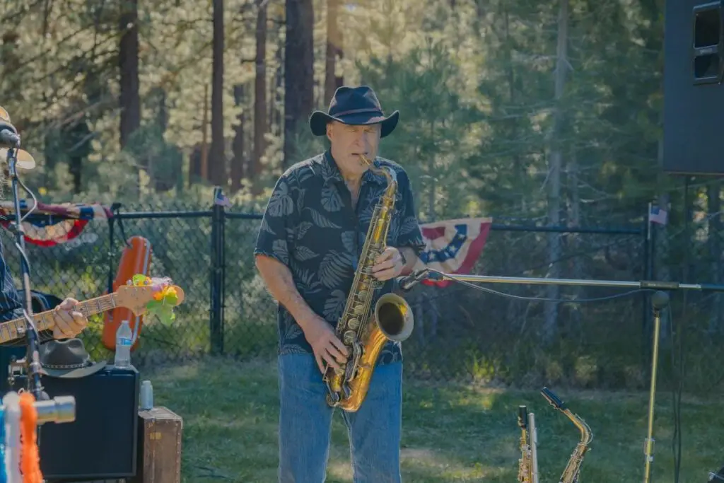 A man wearing a black hat plays a saxophone outdoors, with trees and festive decorations in the background, adding a joyful tune to the Fourth of July celebrations.