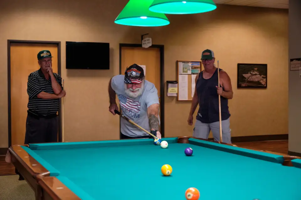 Under the soft glow of green lights in a game room at Gold Canyon RV Resort, three men engage in a friendly game of pool. As one man takes his shot, the others watch intently, enjoying the resort's top-notch amenities during their winter getaway.