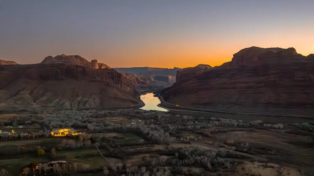 At sunset, the river slices through the canyon, framed by rugged hills and silhouettes. In the foreground, Portal RV Resort's glowing lights illuminate a small settlement, offering a serene retreat amidst nature's grandeur.