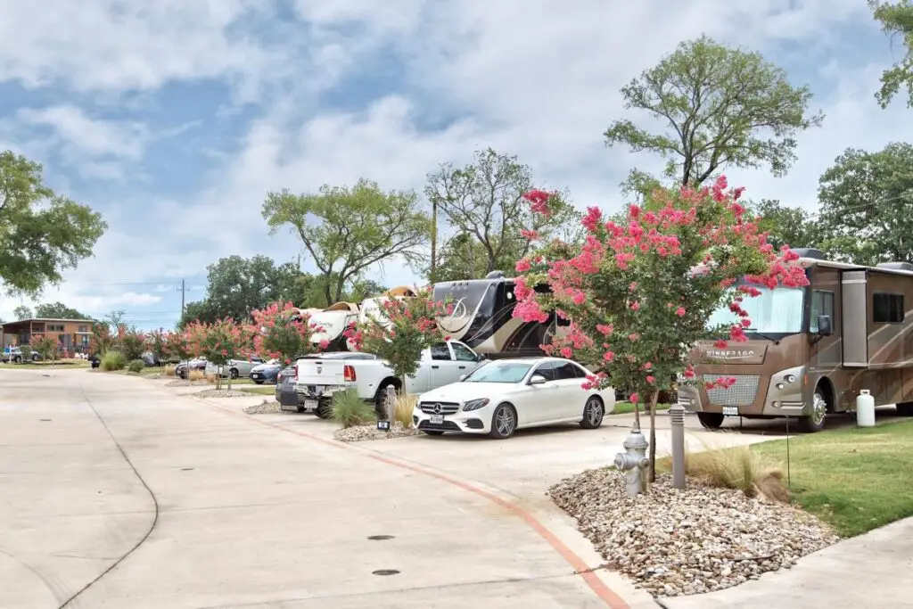 RVs and cars parked along a driveway lined with pink flowering trees and shrubs, reminiscent of an Oak Forest, with a building visible in the background.