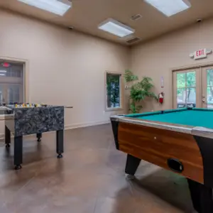 The Rayford Crossing recreation room features a foosball table and pool table, bathed in natural light from large windows, with a wall-mounted TV as the centerpiece.