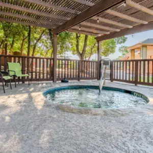 At Rayford Crossing, enjoy a covered outdoor hot tub with bubbling water. Relax in the two green chairs beside a side table, while surrounded by trees and a charming house in the background.