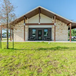The one-story building at Rayford Crossing boasts a stone facade and wooden beams, surrounded by lush grass. Two picnic tables invite visitors to relax, while parked cars can be seen in the background.