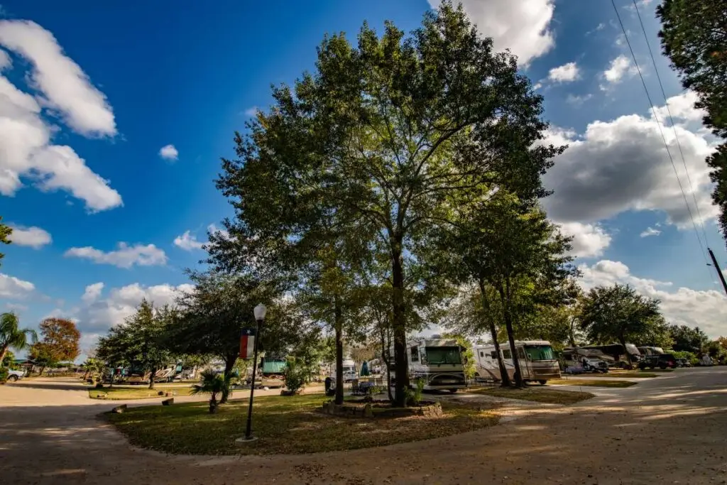 Rayford Crossing RV park features several vehicles parked under large trees, with a clear sky and scattered clouds above. Roads gracefully wind through the serene area.