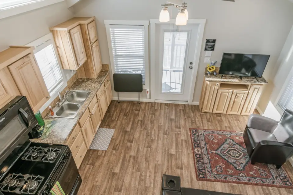A small kitchen and living area with wood cabinets, a cozy Amarillo armchair, and a rug on wood flooring. Natural light flows through windows and glass doors. A stove, sink, TV on a cabinet complete the warm charm typical of Comfortable Cottages.