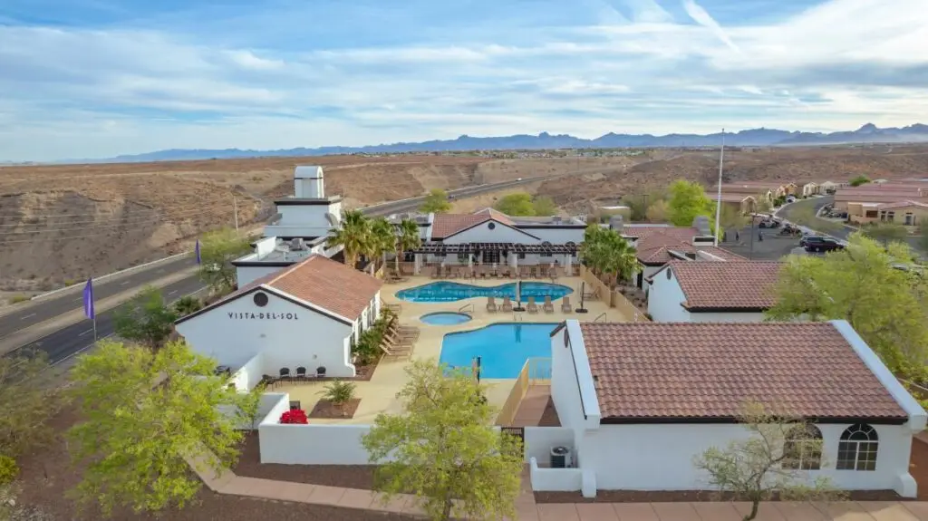 Aerial view of the Vista Del Sol desert community showcases a central swimming pool, surrounded by white buildings with red roofs. A nearby road winds through the scene, while hills rise under a clear sky in the background.