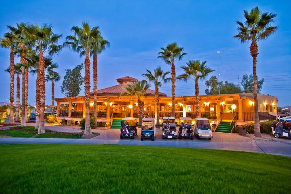 At dusk, the Pueblo El Mirage golf course clubhouse stands elegantly with palm trees swaying gently and parked golf carts lined up in front, all surrounded by meticulously manicured greens.