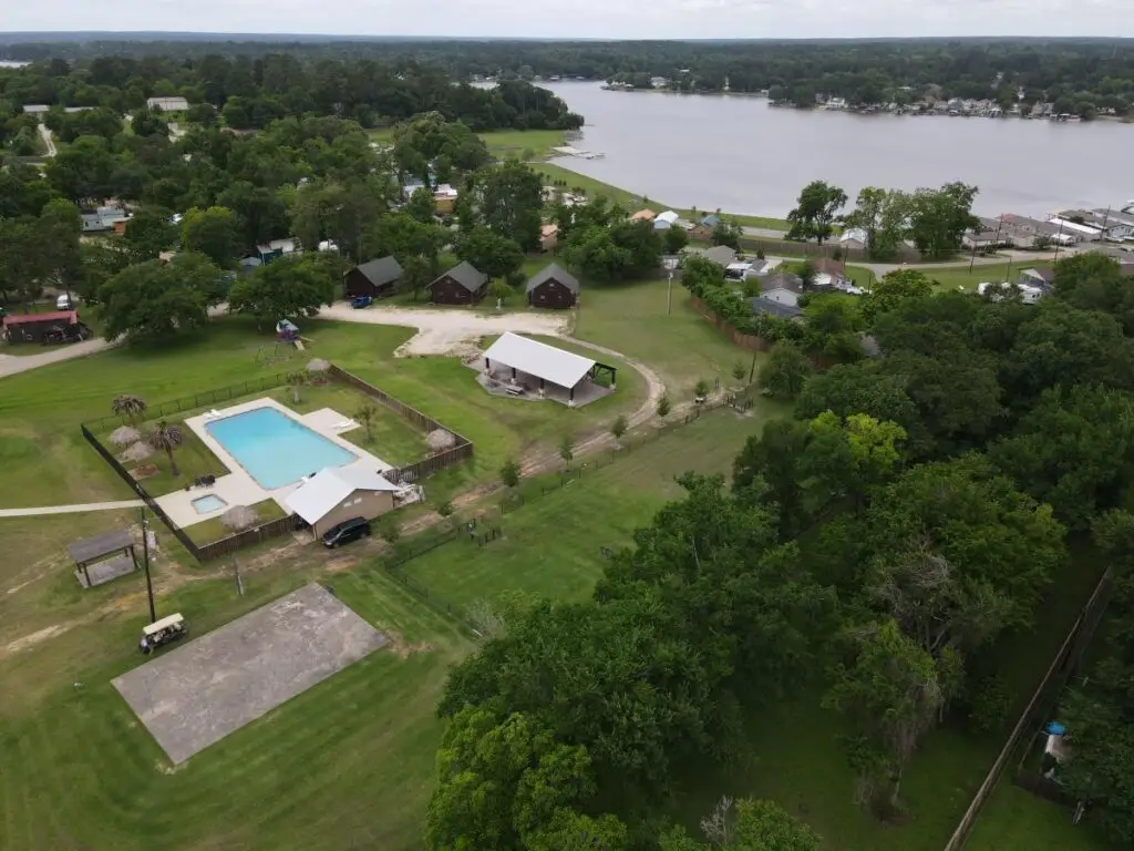 Aerial view of the Water's Edge vacation area with cozy cabins, a sparkling swimming pool, and lush greenery. Lake Conroe completes the stunning backdrop, offering a picturesque retreat.