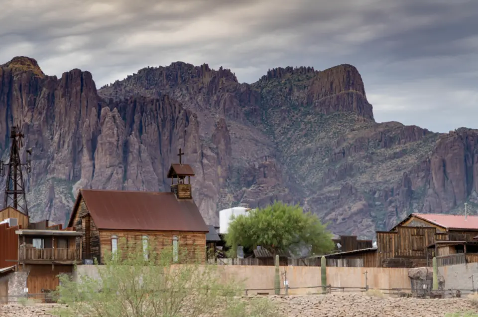 Set against a backdrop of rugged, rocky mountains under a cloudy sky, the old western town, nestled near Gold Canyon, features wooden buildings and a classic church. Book your RV site today to experience the charm and history of this enchanting destination.