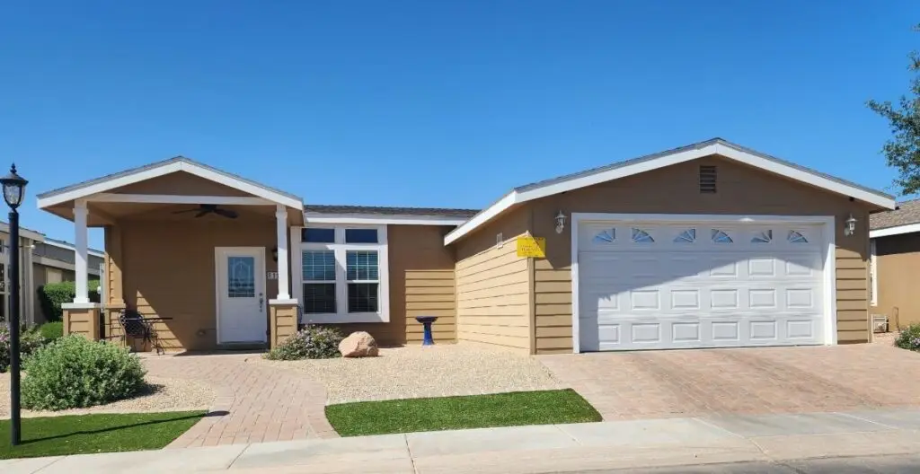 The F119 single-story house features brown siding, a welcoming front porch, a white door, and a double garage. The yard offers minimal landscaping with gravel and artificial grass under a clear blue sky.