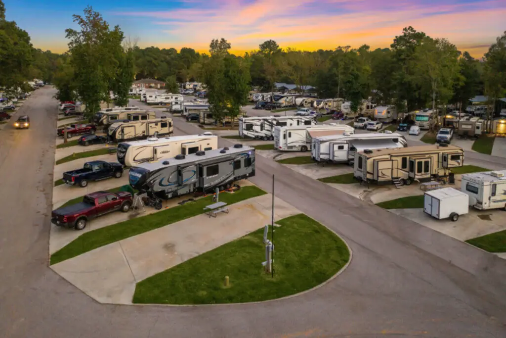 A stunning aerial view of an RV campground at sunset reveals several parked RVs and trucks on paved lots, all surrounded by lush trees.