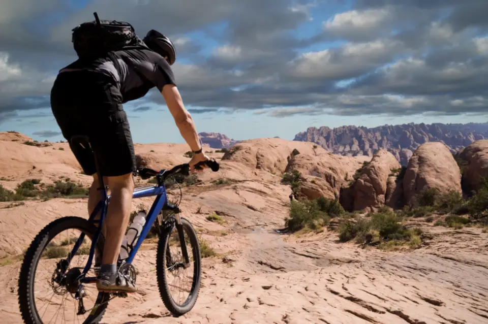 A person is mountain biking on a rocky trail with a backpack under the expansive blue sky and fluffy clouds. This adventurous ride feels like the perfect Father's Day escape, embracing nature's challenges and exhilaration.