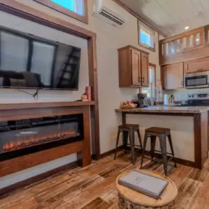 Modern tiny house interior perfect for vacation rentals, featuring a wall-mounted TV, an electric fireplace, and a kitchen area with cabinets, countertop stools, and stainless steel appliances.