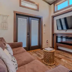 Cozy living room in this vacation rental features a brown sofa, wall-mounted TV above an electric fireplace, and a round wicker stool. Large glass double doors and windows flood the space with natural light.