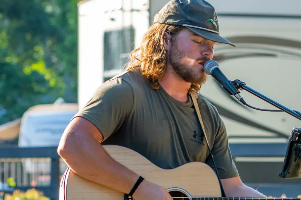 A man with long hair and a cap plays an acoustic guitar and sings into a microphone outdoors, serenading adventurers as they embark on nearby hiking trails.