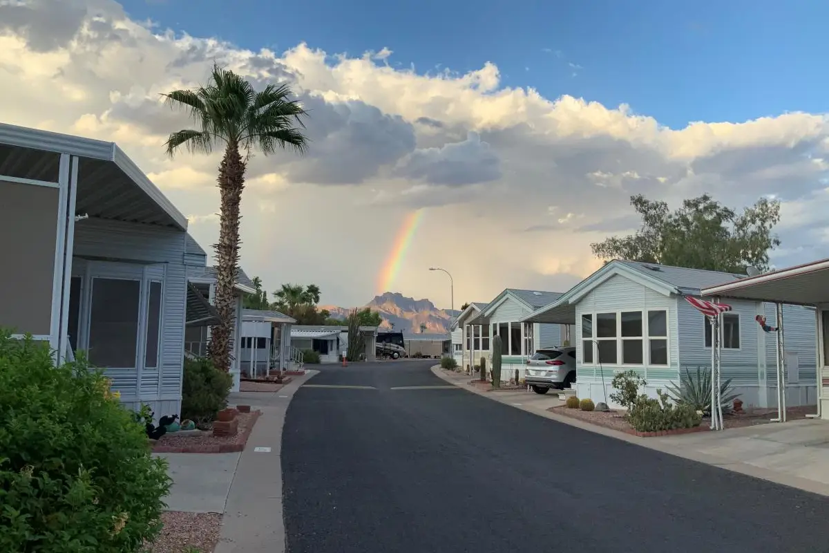 A residential street lined with manufactured homes, palm trees, and desert landscaping transforms into a picturesque setting as a rainbow arches across the cloudy sky, mountains standing majestically in the background—an idyllic scene for potential vacation rentals.