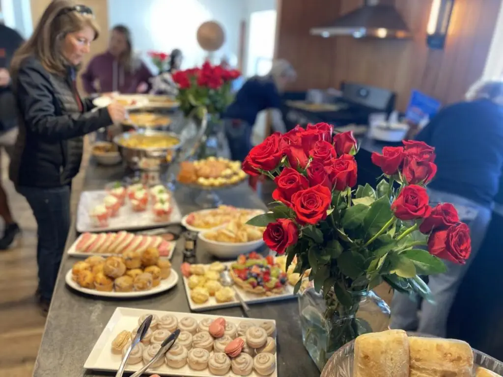 A buffet table adorned with assorted pastries, cookies, and sandwiches sits elegantly amidst a bouquet of red roses, creating May Memories! Families and friends are serving themselves in the background, cherishing the moment.