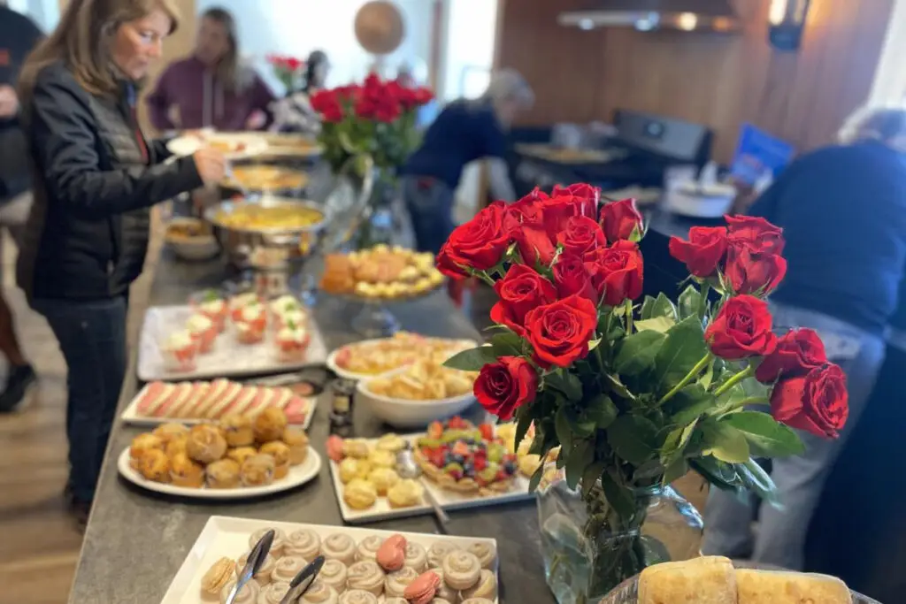 A buffet table brimming with pastries, skewers, and desserts is adorned with red roses. As people serve themselves in the background, this scene captures the essence of creating memorable experiences.