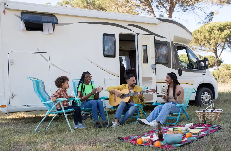 A family enjoys a sunny day outside their camper van at Pueblo El Mirage. Two children clap along as a man strums his guitar, while a woman smiles warmly. A picnic blanket with an array of treats is spread on the grass, capturing the spirit of this 55+ resort getaway.