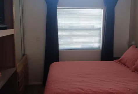 A small 2010-themed bedroom features a pink bedspread, dark curtains, a window with blinds, and a wooden dresser.