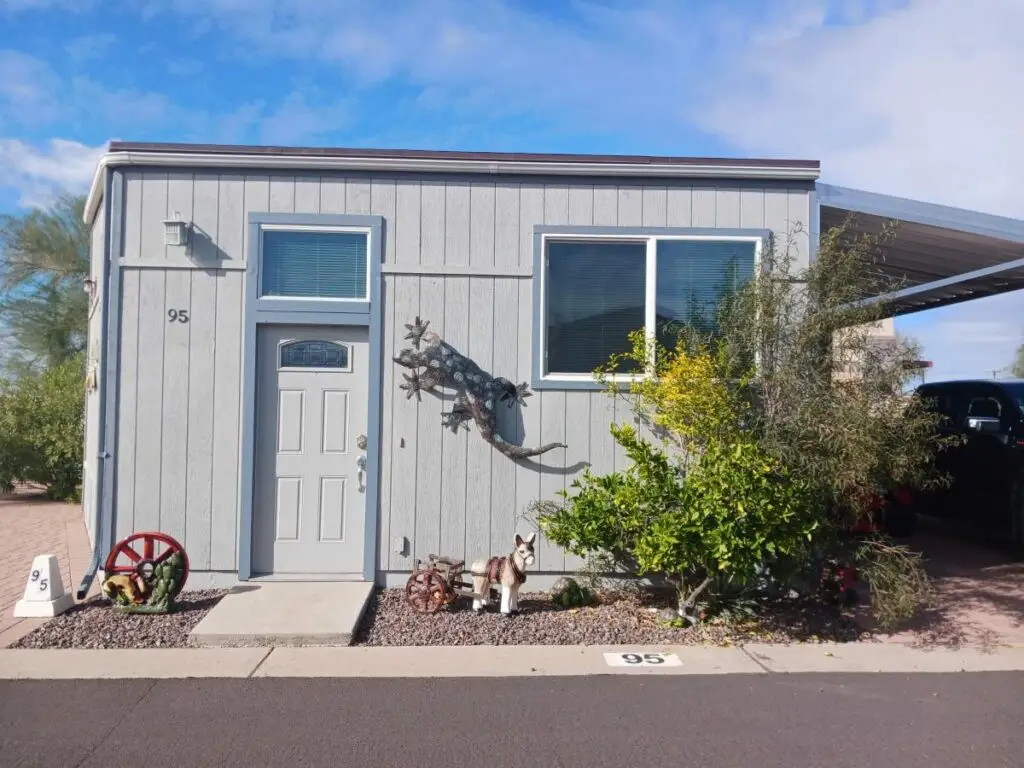 A charming mobile home, reminiscent of a 2010 Chariot Eagle, features a decorative metal lizard on its wall. Two red wagon wheels and vibrant plants add flair. A figure of a dog sits on the gravel by the door, under house number 95.