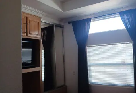 The cozy mobile home bedroom features a bed, large window with curtains, a wardrobe, and a built-in TV. Natural light beautifully illuminates the space.