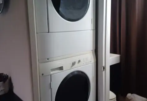 In this stylish Chariot Eagle manufactured home, you'll find a stacked washer and dryer conveniently placed next to a half-open door, unveiling a bathroom adorned with a dark curtain and pristine white sink.