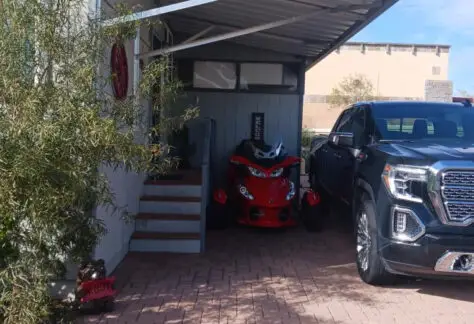 A red motorcycle and a black pickup truck are parked under a metal carport beside a Chariot Eagle RV. A plant is near the driveway, adding a touch of greenery to the scene.