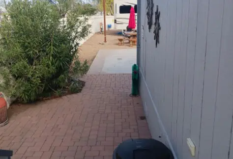 Paved walkway alongside a building, with a trash can in the foreground, leads to a gravel area featuring a picnic table and a Chariot Eagle mobile home from 2010 in the background. A bush is on the left.