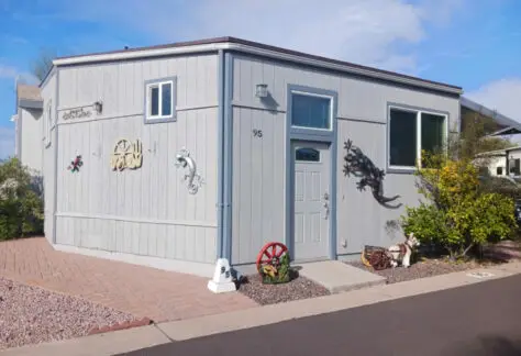 A small, modern gray building resembling a 2010 Chariot Eagle RV with decorative wall art and a white front door labeled "#95" sits on a paved walkway surrounded by gravel and minimal landscaping.