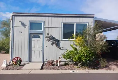 A charming mobile home, reminiscent of a 2010 Chariot Eagle, features a decorative metal lizard on its wall. Two red wagon wheels and vibrant plants add flair. A figure of a dog sits on the gravel by the door, under house number 95.