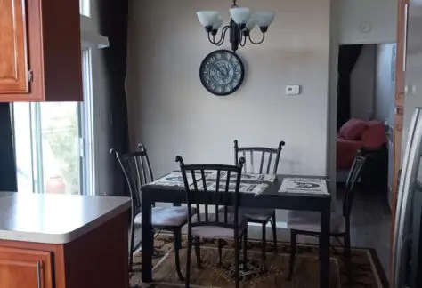 The dining area, reminiscent of a sleek Chariot Eagle design, features a black table with four chairs, a chandelier above, and a clock on the wall. This stylish space seamlessly opens to the living area where you catch a glimpse of the inviting red sofa.