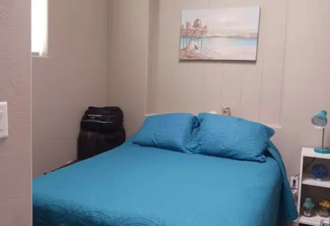 A cozy small bedroom featuring a Chariot Eagle vibe: a turquoise blanket draped over the bed, a suitcase tucked in the corner, a nightstand adorned with lamps and decor, and an elegant painting above the headboard.