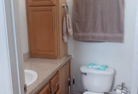 This compact bathroom, reminiscent of a 2010 Chariot Eagle mobile home, features a toilet, wooden cabinets, countertop, and sink. A brown towel elegantly drapes above the toilet, and a pack of wipes rests conveniently on the tank.