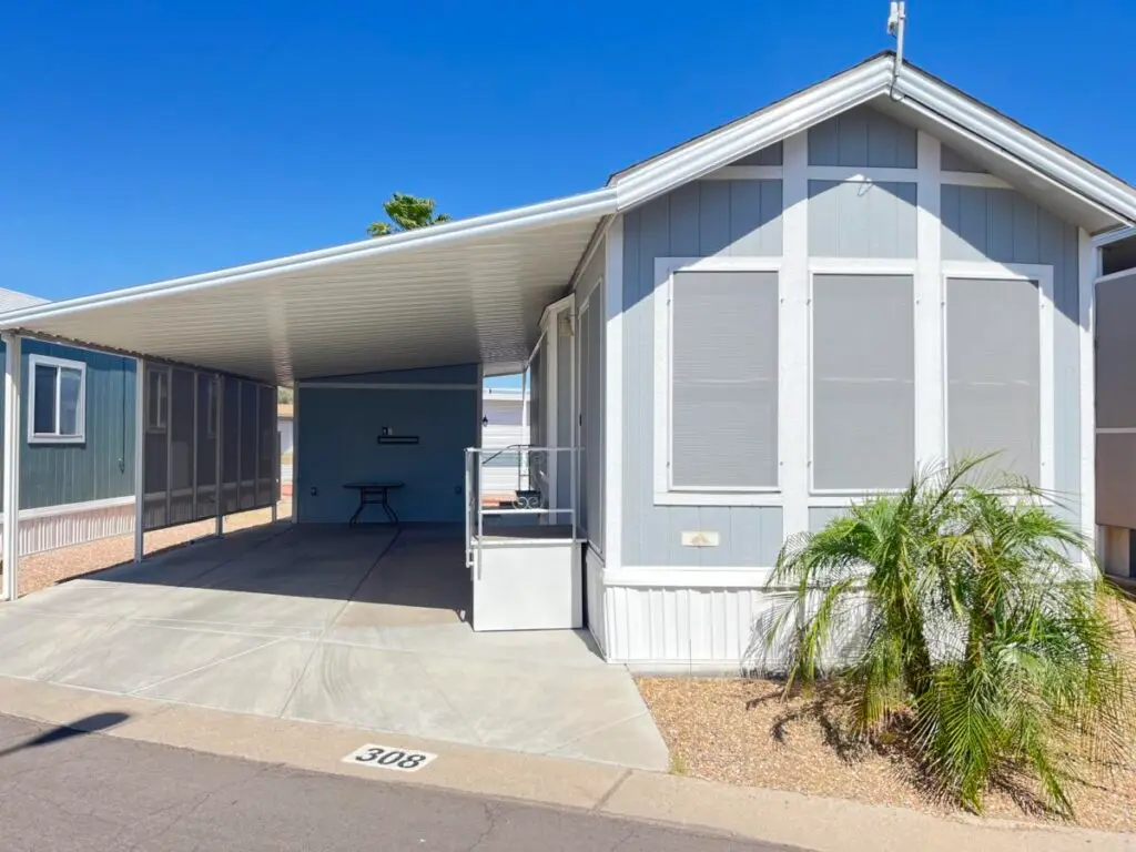 A gray 2015 Cavco mobile home with white trim features a covered carport and stairs. A small palm plant adorns the front, while another home is visible on the left, subtly marked as #308.