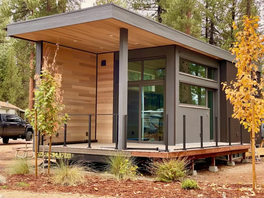 A modern tiny house with a wood and glass exterior sits like an adventure cabin on a raised platform, surrounded by trees and plants in a serene natural setting.