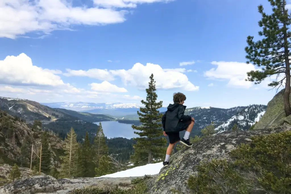 A person climbs a rocky hill in Coachland, overlooking a scenic view of a lake, mountains, and pine trees under a partly cloudy sky.