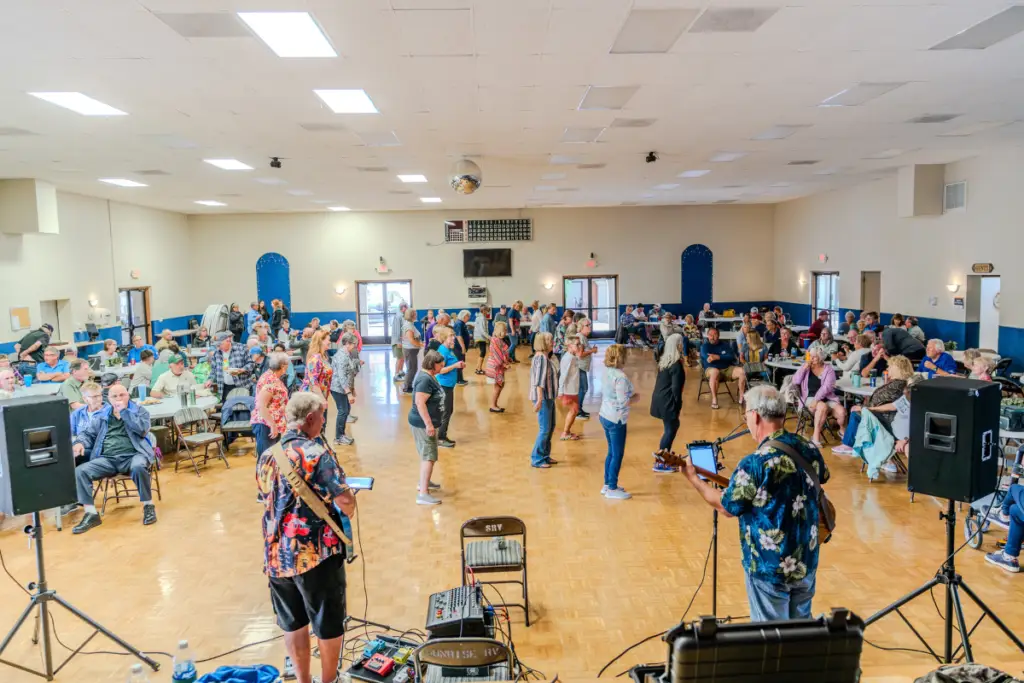 In the vibrant community hall of Sunrise RV Resort, a band performs as attendees explore the lively atmosphere, mingling and dancing amidst the amenities.