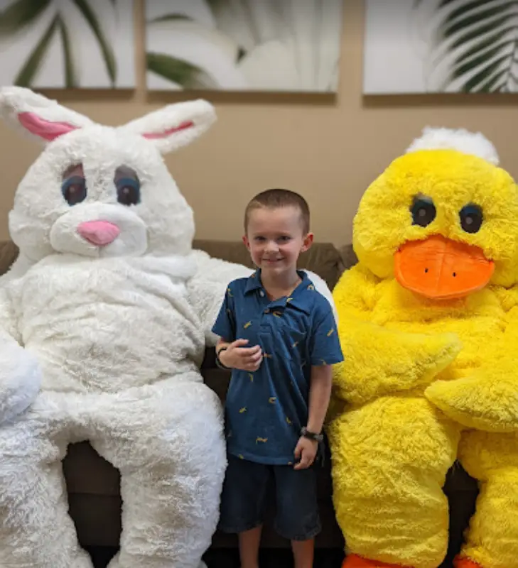 A child sits between a person in a white bunny costume and a person in a yellow duck costume, all nestled on a cozy couch at an Easter event.