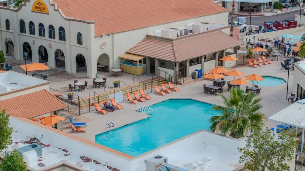 Aerial view of a hotel courtyard at sunrise, featuring two swimming pools, orange lounge chairs, and umbrellas. Guests are enjoying relaxation as they lounge around the pools.