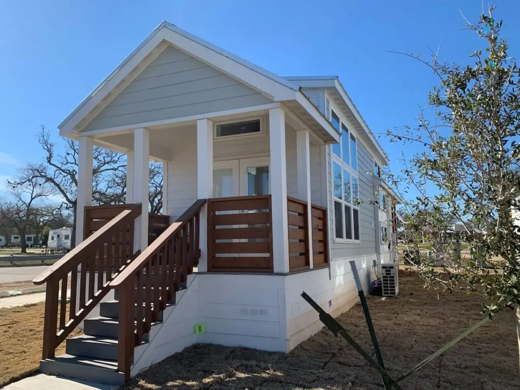 A small white house with a wooden porch and staircase, reminiscent of a Bergstrom retreat, sits on a grassy lot under a clear blue sky.